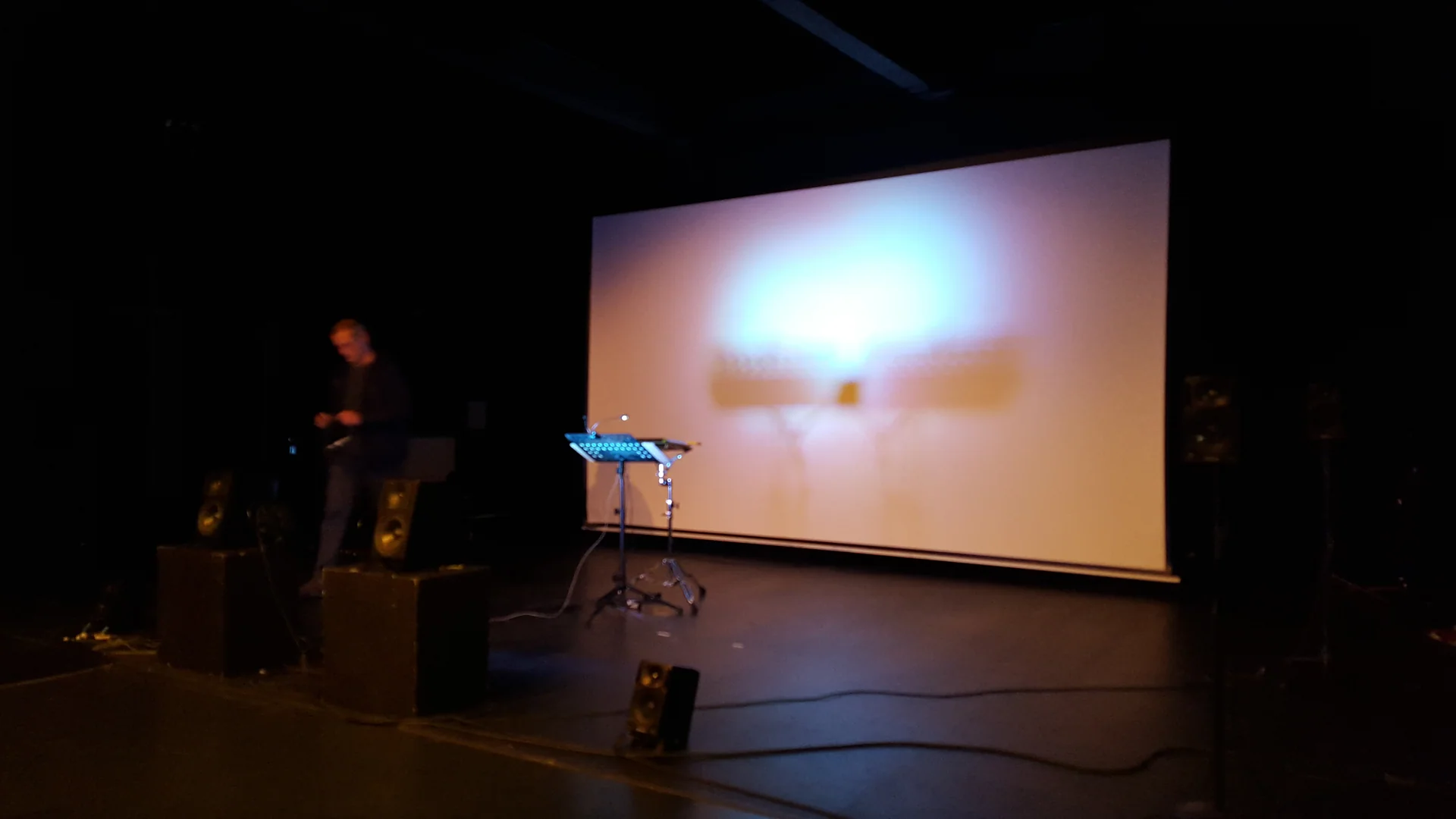 A performer, speakers and an electronic percussion setup in front of a projection screen